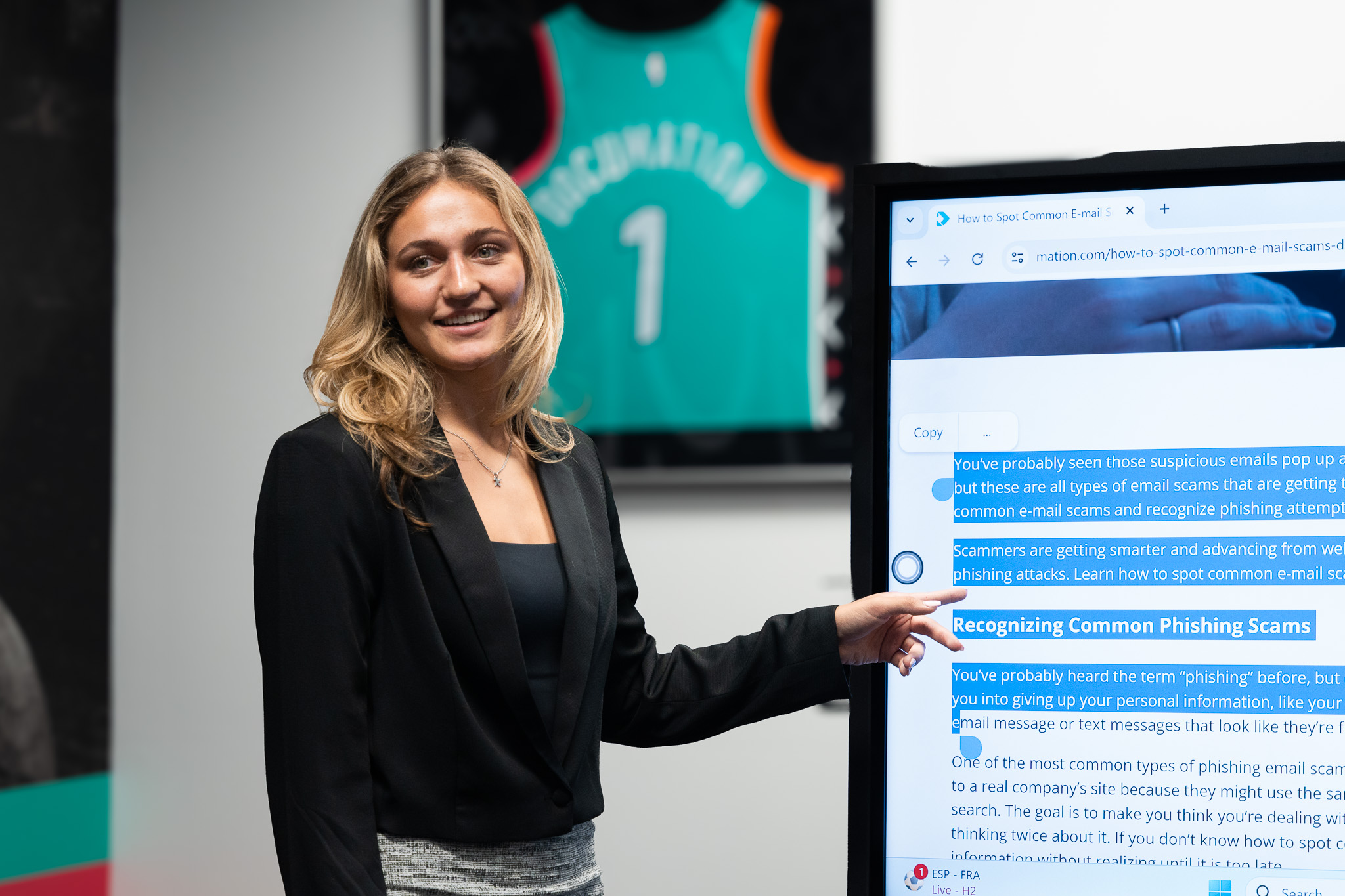A woman with blonde hair is standing next to an interactive display screen, smiling and pointing to text about recognizing common phishing scams. She is wearing a black blazer and gray skirt. In the background, a teal San Antonio Spurs jersey with 