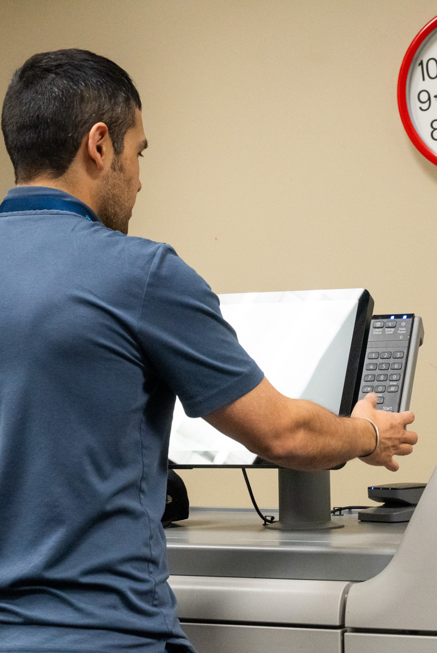 Man operating a touchscreen control panel on a large multifunction printer or copier in an office setting.