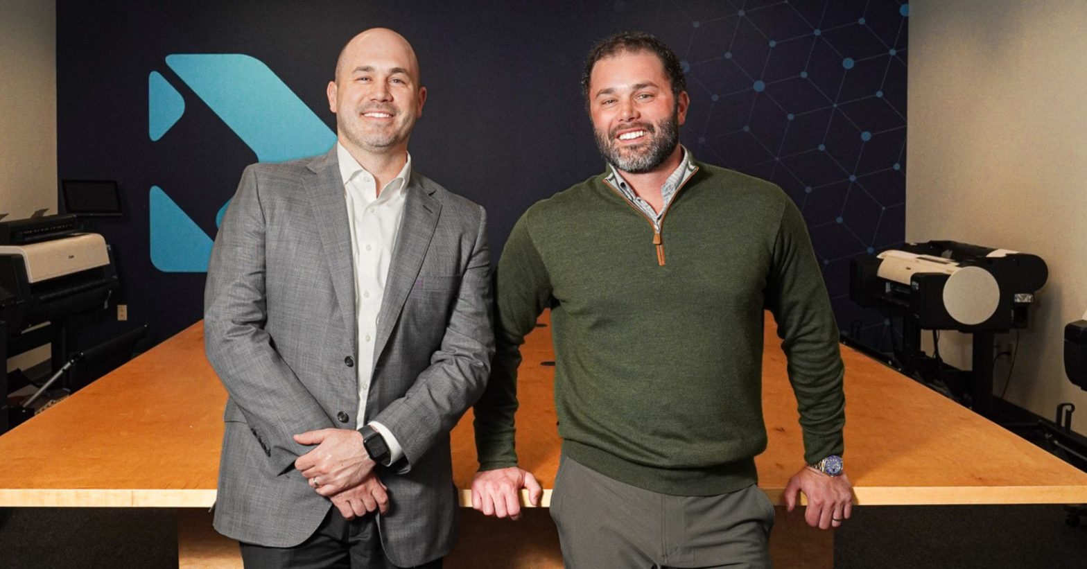Preston Woolfolk and Hunter Woolfolk, co-CEOs of DOCUmation, stand side by side in the company's demo room. Preston is dressed in a gray suit, while Hunter is wearing a green sweater.