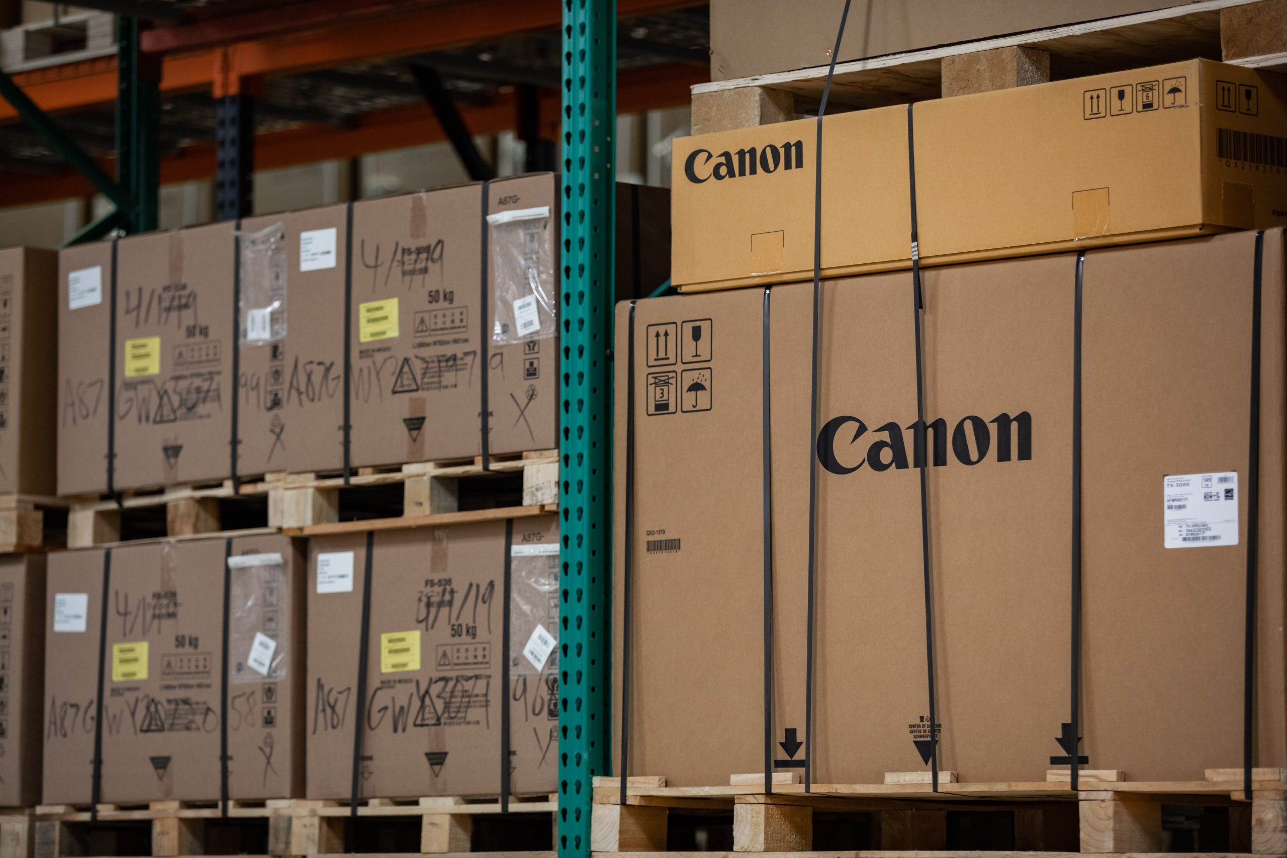 Stacked pallets of Canon office equipment boxes in a warehouse, secured with straps.