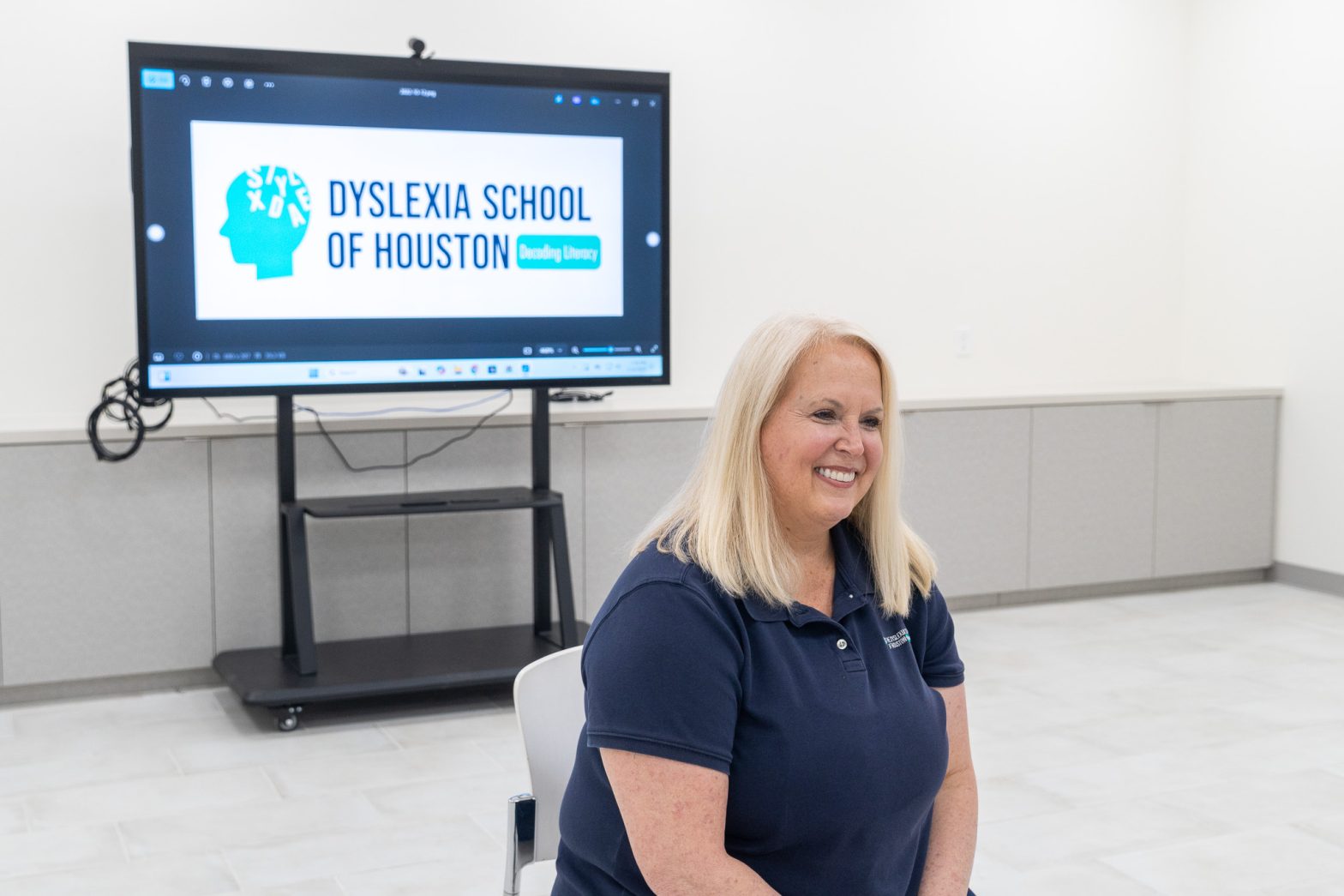 Tammy Spencer, founder and CEO of the Dyslexia School of Houston, smiles while sitting in a modern classroom. Behind her, a large screen displays the school's logo with the tagline "Decoding Literacy."