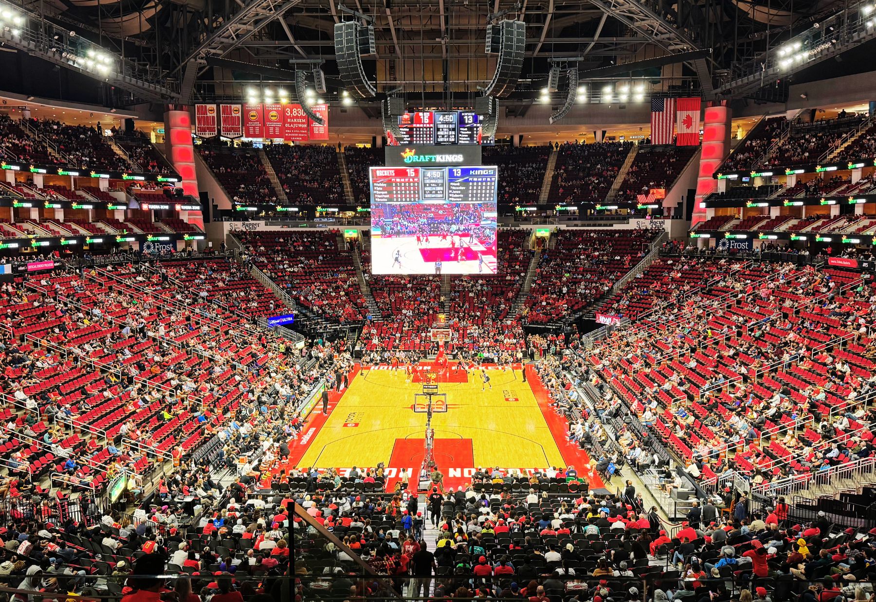 Stadium full of with fans watching a basketball game at the Houston Rockets' arena. The court is brightly lit, surrounded by red seating, and players can be seen in action.