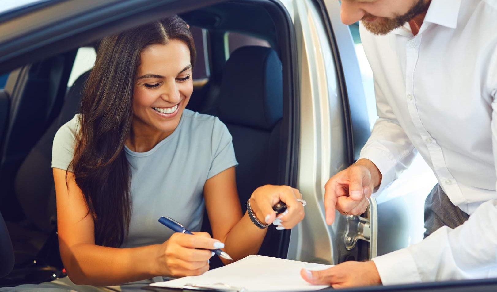 A smiling woman sits in a car while signing paperwork, holding car keys, as a salesperson in a white shirt assists her.