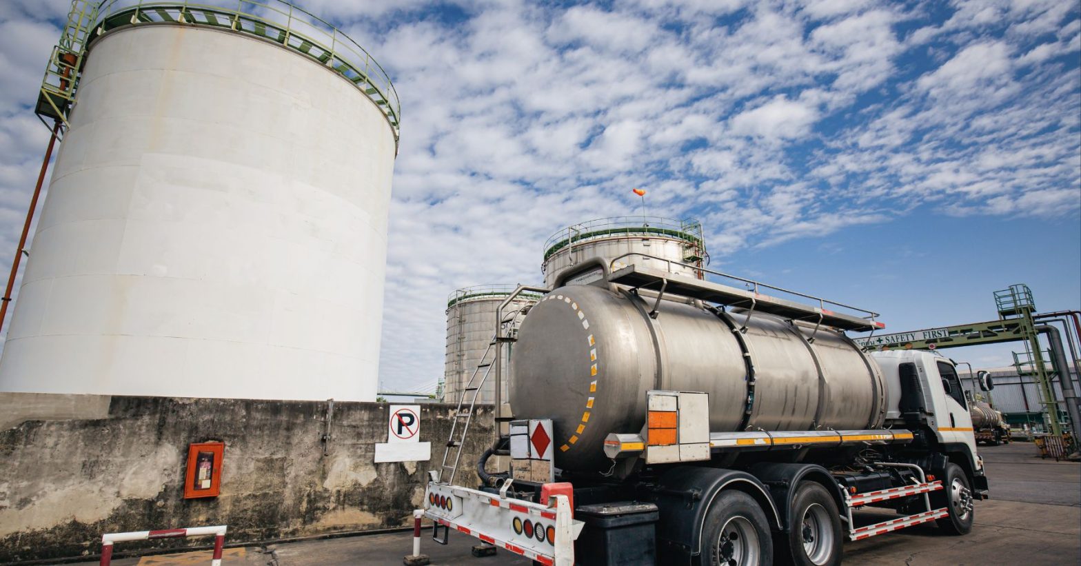 A fuel tanker truck parked near large industrial storage tanks at a processing facility under a blue sky.