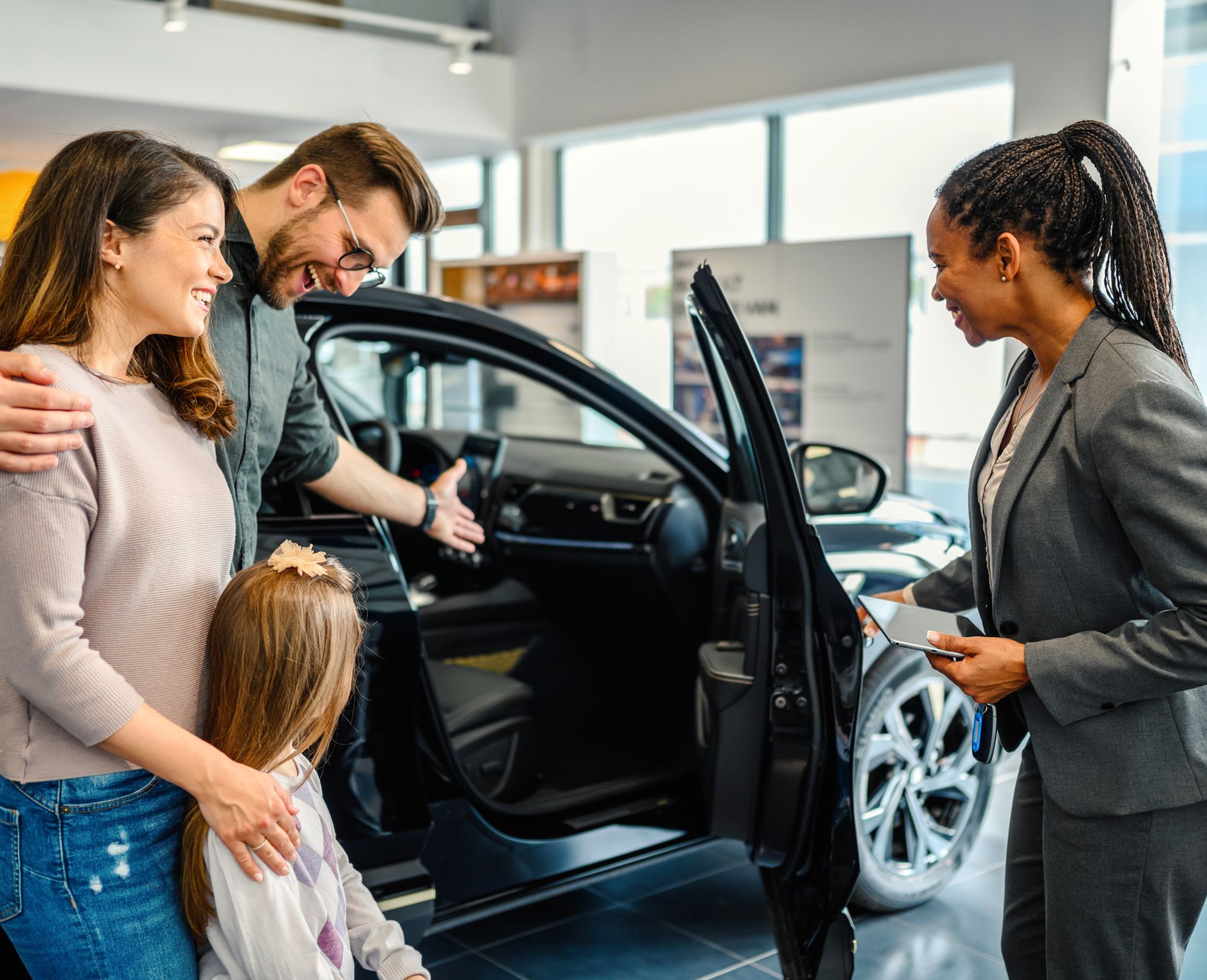 A smiling family explores a new car with the help of a dealership sales representative in a showroom.