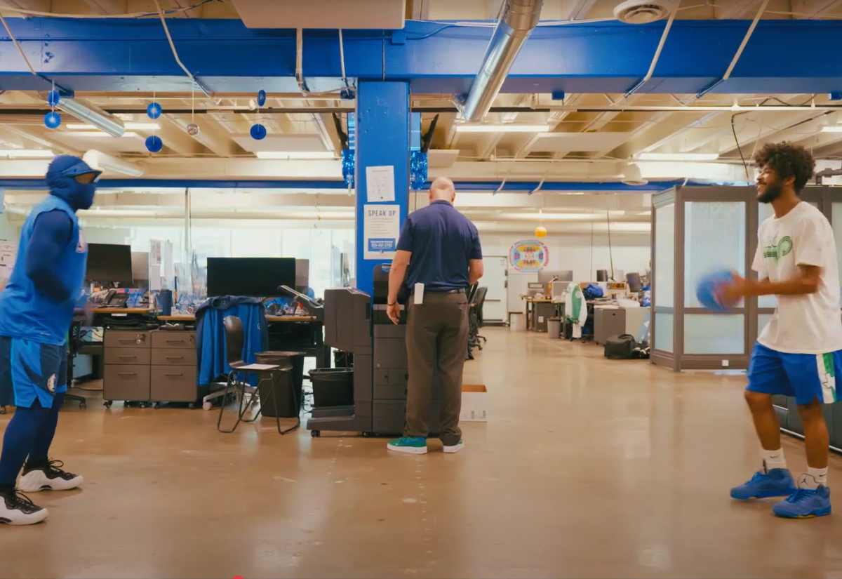 MavsMan, the Dallas Mavericks mascot, dressed in a blue full-body suit and basketball gear, plays with a staff member holding a blue ball inside the Dallas Mavericks HQ. A man in a navy blue polo and brown pants stands at a copier, seemingly unaware of the activity behind him. The office environment has blue decorations, desks, computers, and an open workspace setup.