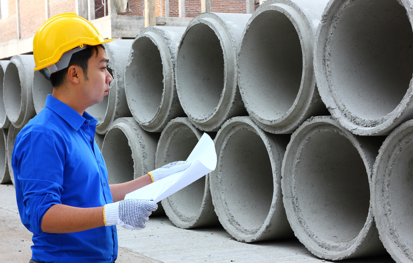 Construction engineer wearing a yellow hard hat inspects blueprints in front of stacked concrete pipes at a job site.
