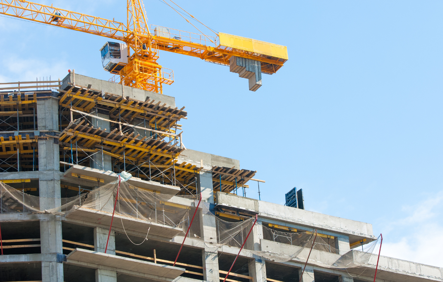 A high-rise building under construction with scaffolding, safety nets, and a large yellow tower crane against a clear blue sky.