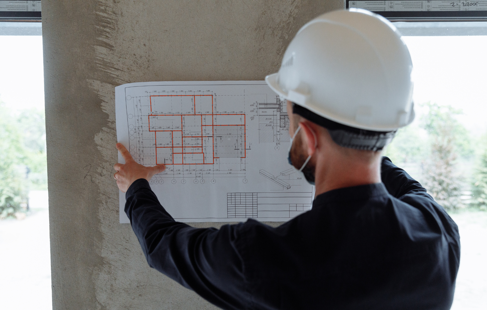 Construction professional wearing a white hard hat reviews and points to a detailed building blueprint taped to a concrete wall.