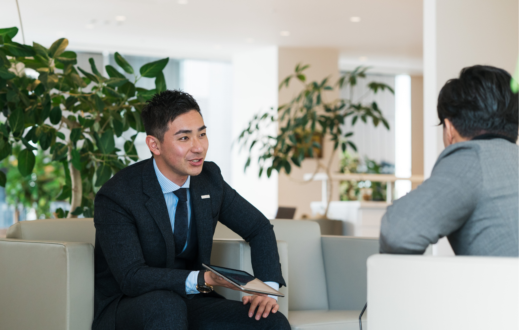 A business professional in a dark suit holds a tablet and engages in a conversation with a client in a modern office setting.