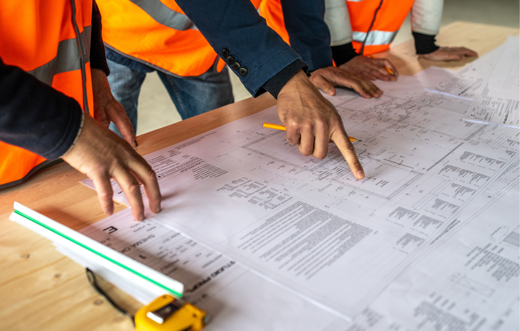 Construction professionals wearing orange safety vests review detailed blueprints on a wooden table, pointing at key project details.