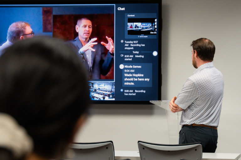 Man at a clear podium interacting with a large interactive screen showing a video conference with speakers, chat messages, and multiple meeting views.
