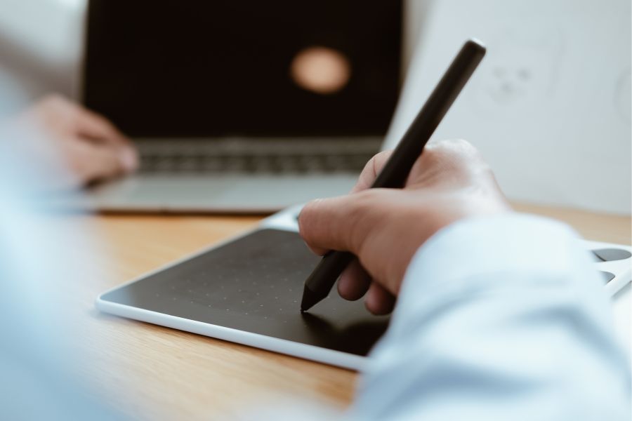 Close-up of a person using a digital drawing tablet with a stylus, sketching near a laptop—ideal for creative design work or preparing artwork for Risograph printing.