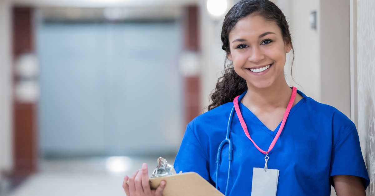 Smiling healthcare worker in blue scrubs holding a clipboard and wearing a stethoscope in a hospital hallway.