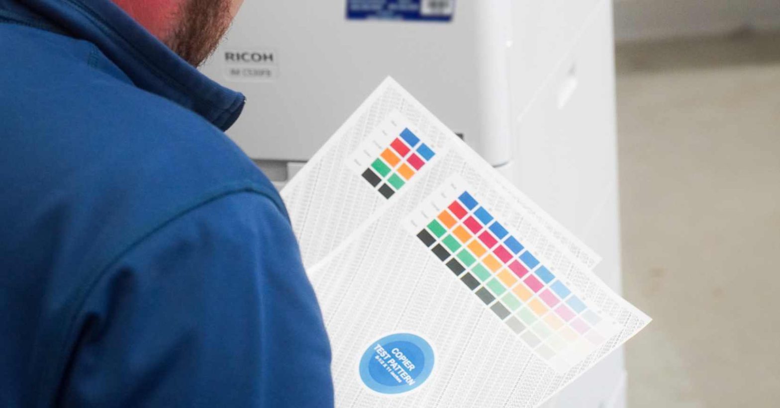 DOCUmation copier technician in a blue jacket holding two color test pattern sheets in front of a Ricoh printer during a print quality check.