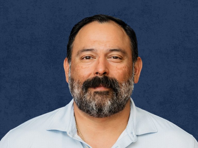 Professional headshot of a man with dark hair and a full salt-and-pepper beard, photographed against a blue textured background.