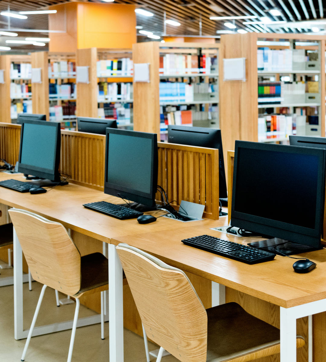 A row of desktop computers with keyboards and mice on wooden desks in a modern library setting.
