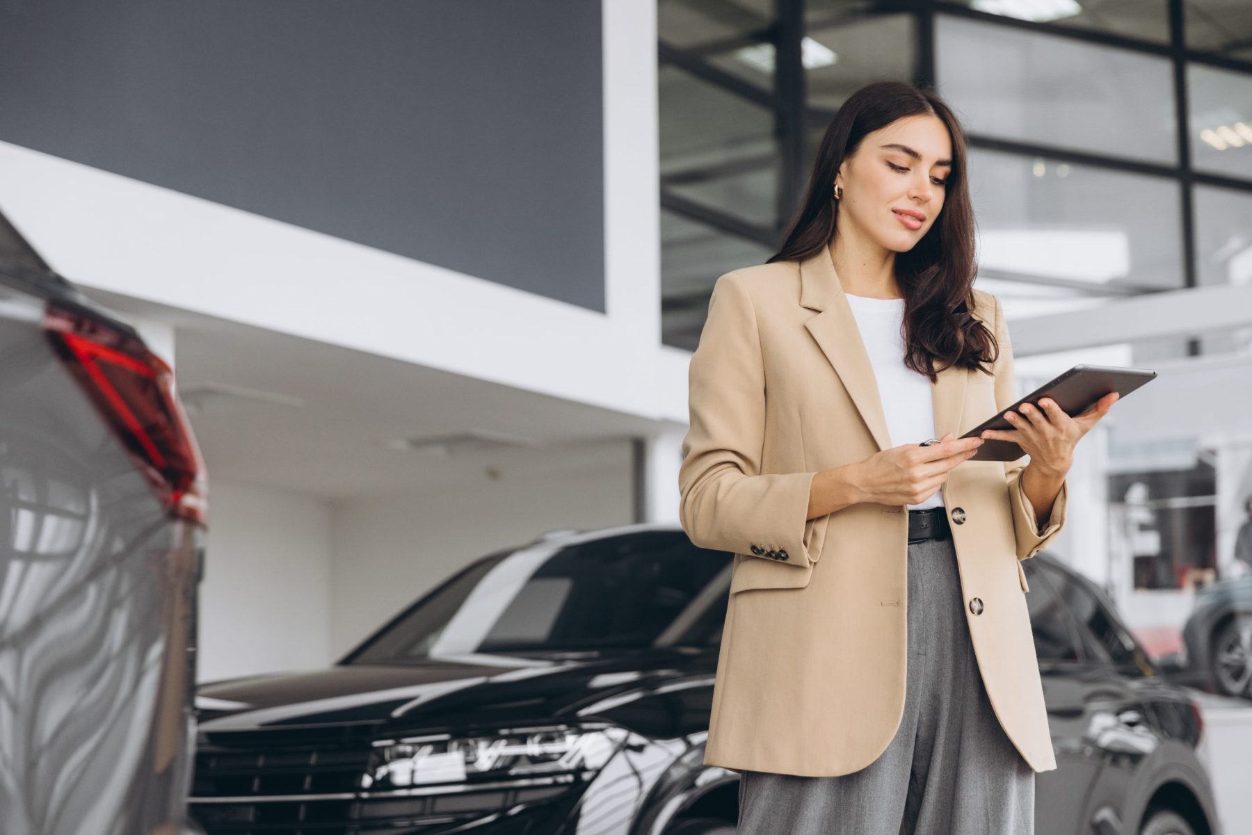 Woman in a tan blazer using a tablet in a modern car dealership showroom beside parked vehicles.