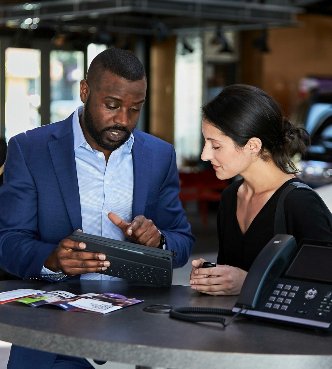 Business consultant demonstrating a tablet and VoIP phone system to a client in a modern automotive showroom.